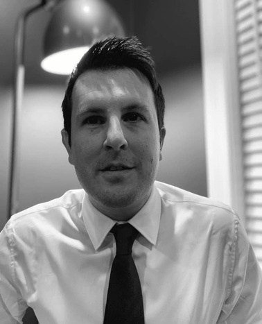 A black and white photo of a man in a white shirt and tie sitting in an office environment.