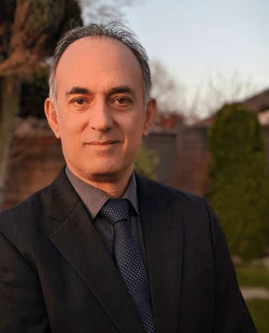 A middle-aged man in a suit stands outside with a soft focus background of greenery during sunset.