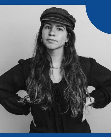 A woman wearing a cap and a black blouse poses against a plain background, with her hands on her hips.
