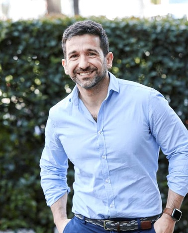 A smiling man standing outdoors wearing a blue shirt.