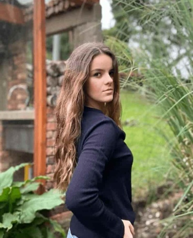A young woman standing outside with trees in the background, looking over her shoulder.