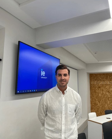 A man in a white shirt standing in a conference room with a large screen displaying 'IE University'.