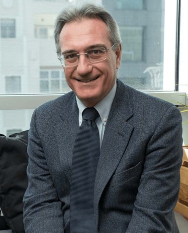 A middle-aged man smiling, wearing a grey suit and glasses, sitting indoors with a window background.