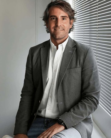 A man in a gray blazer and white shirt sits by a window with partially closed blinds, smiling subtly towards the camera.