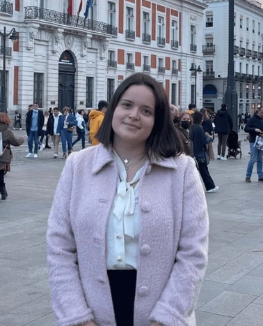 A young woman in a pink coat and white blouse standing in front of a historic building with people in the background.