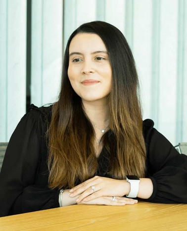 A woman sitting at a table with a confident smile, wearing a black blouse and a smartwatch.