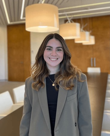 A smiling young woman wearing a grey blazer and black top, standing in an office with modern hanging lamps and wooden paneling in the background.