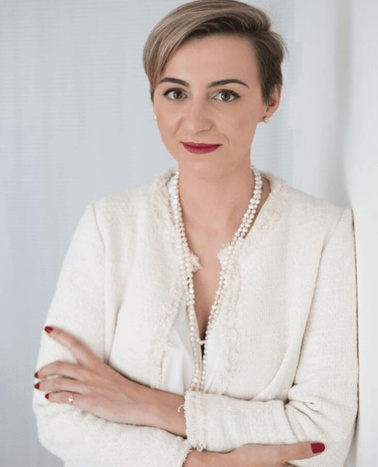 A professional woman in a white jacket and pearl necklace is posing with crossed arms against a light background.