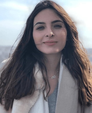 A woman with long brown hair wearing a white coat and standing outdoors.