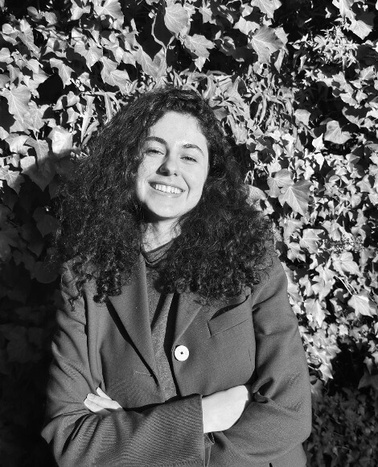 A black and white photo of a smiling woman with curly hair, standing in front of a leafy background.