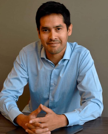 A man in a light blue shirt sitting at a table with folded hands, smiling at the camera.