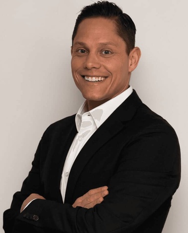 A smiling man in a black suit and white shirt posing against a light grey background.