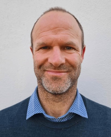 A smiling bald man wearing a blue sweater and a checkered shirt standing against a white background.