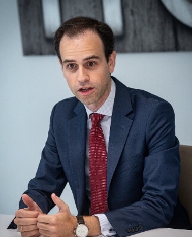 A man in a formal blue suit and red tie sitting at a table during a meeting.