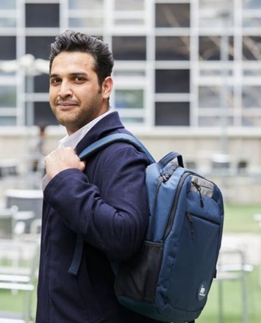 A confident man in a suit jacket with a backpack standing in front of a building with a grid-like structure.