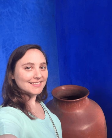 A woman smiling next to a large terracotta pot against a vivid blue wall.