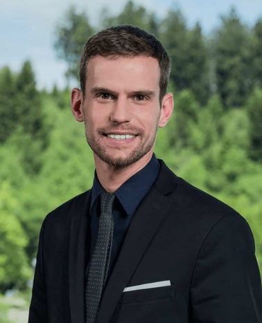 A professional man in a suit smiling against a blurred natural background.