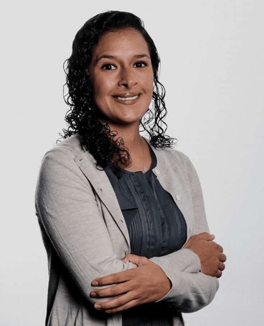 Portrait of a smiling woman with curly hair, wearing a gray blazer over a blue blouse, standing with her arms crossed.