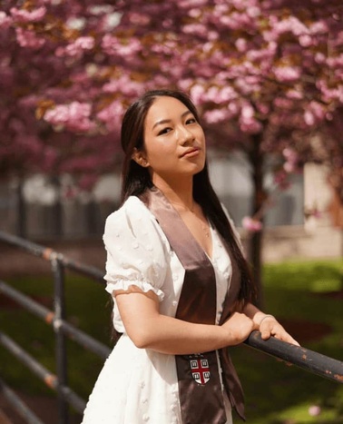 A young woman in a white dress stands by a railing, with blooming pink cherry blossoms in the background.