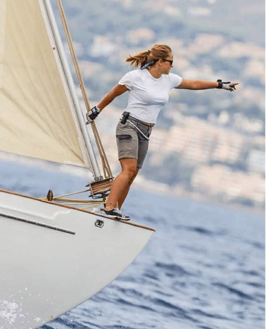 A woman balances on the edge of a sailing boat, extending her arms for stability.