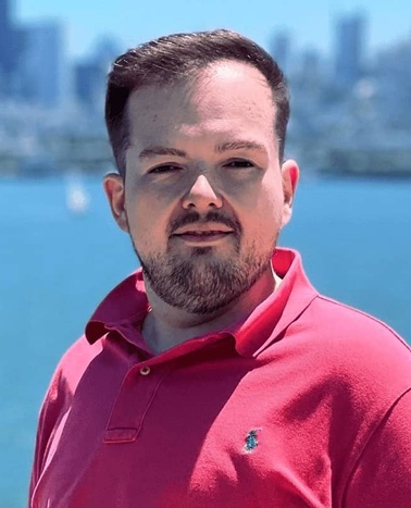 A man in a red polo shirt standing before a body of water with a city skyline in the background.