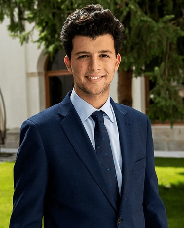 A young adult male in a blue suit and tie standing in front of a building with trees in the background.