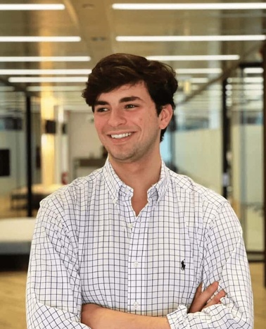 A young man smiles while standing in an office environment, wearing a checkered shirt with arms crossed.