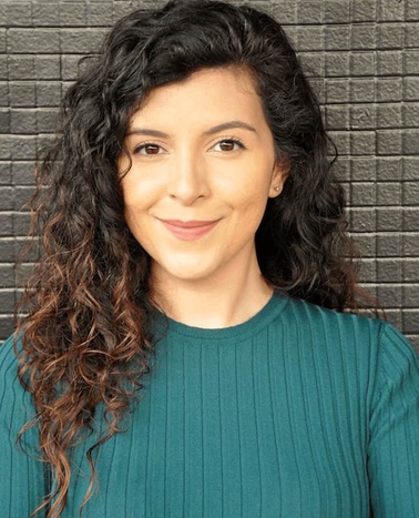 A woman with curly hair smiling at the camera against a brick wall background.