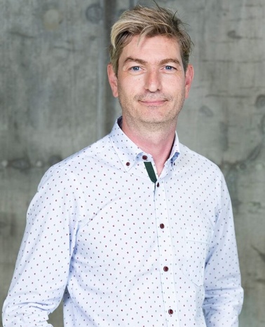 A man in a dotted shirt smiling at the camera with a concrete wall in the background.