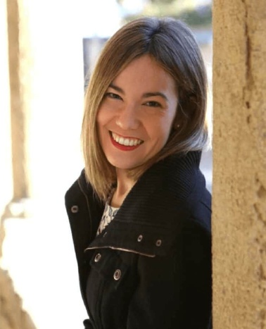 A smiling woman with blonde hair, wearing a black jacket, leaning against a stone pillar.