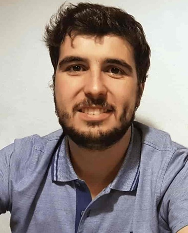 A portrait of a smiling young man with dark hair and a beard, wearing a blue polo shirt against a white background.