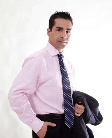 A man in a pink shirt and blue tie holding a jacket over his shoulder poses against a white background.