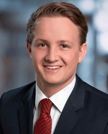 A professional headshot of a young man with blonde hair, smiling, wearing a dark suit and red tie.