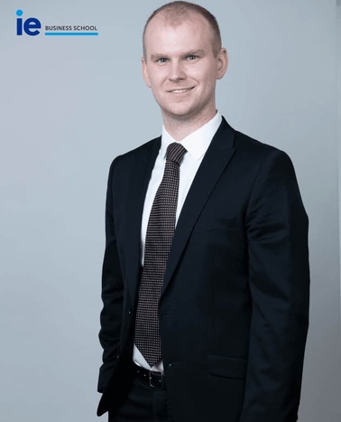 A professional portrait of a man in a business suit with a confident smile, standing against a grey background.