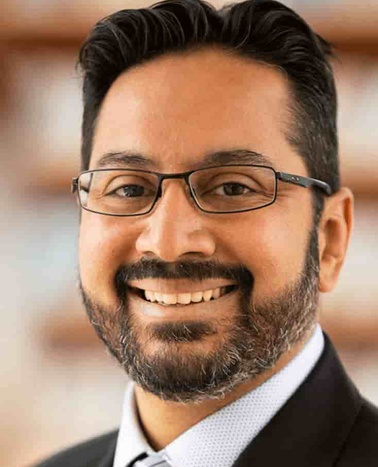 Portrait of a smiling man with a beard wearing glasses and a suit.