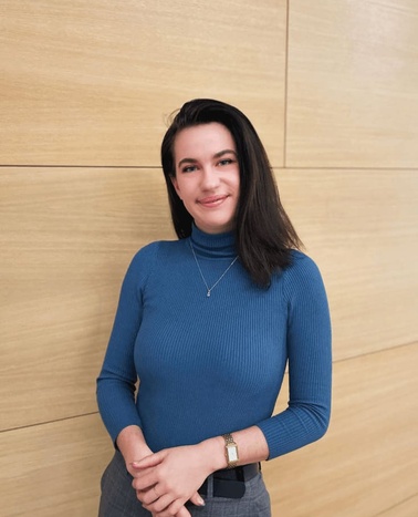 A woman in a blue turtleneck stands confidently with her hands clasped in front of a wooden wall.