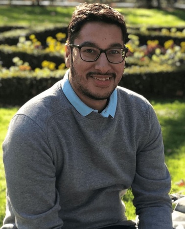 A smiling man wearing glasses, a sweater over a collared shirt, seated outdoors with flowers and greenery in the background.