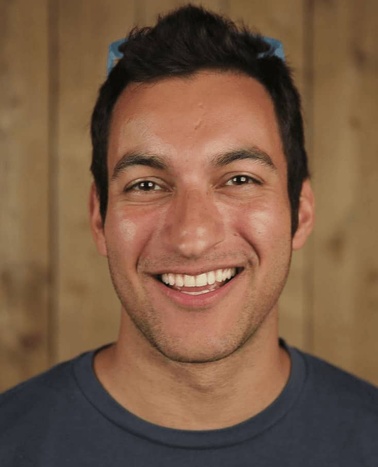 A portrait of a smiling man with dark hair wearing a blue t-shirt, set against a wooden background.