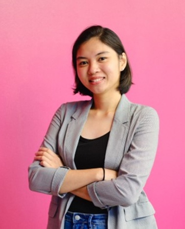 A young woman in a gray blazer and jeans stands smiling with her arms crossed against a pink background.