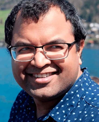A smiling man with glasses wearing a blue polka dot shirt, with a blue water body and hills in the background.