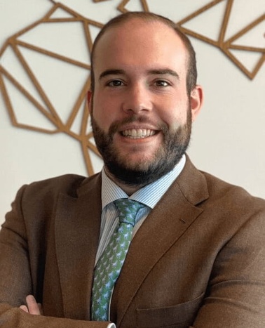 A professional portrait of a smiling man in a brown suit with a blue tie, standing in front of a geometric wall art.