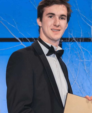 A young man in a tuxedo holding a certificate at an event with a blue background.