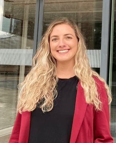 A smiling woman with curly blonde hair, wearing a black top and a red blazer, standing in front of a glass door.