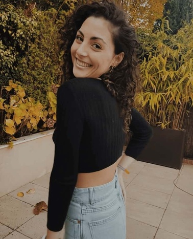 A smiling woman with curly hair, wearing a black top and blue jeans, stands in a garden.