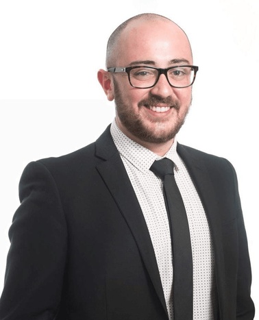 A bald man with glasses wearing a black suit and patterned tie smiling against a white background.