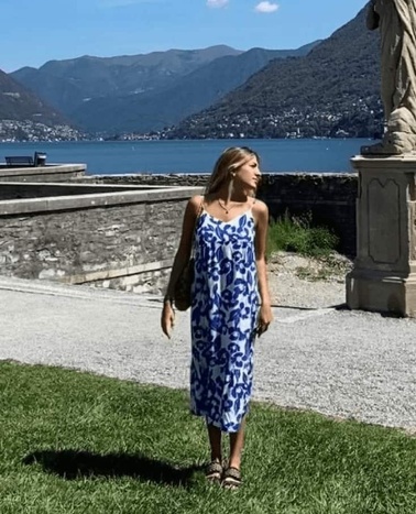 A woman in a blue and white dress walking by a lakeside with mountains in the background.