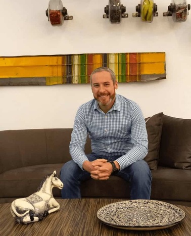 A smiling man in a blue checked shirt sitting on a dark sofa in a room with decorative items on the wall and on the table.