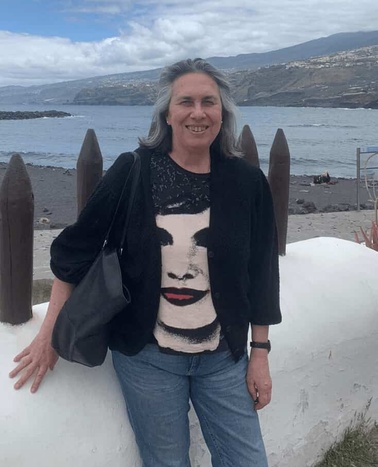 A middle-aged woman with long grey hair smiling, wearing a black cardigan and a t-shirt with a face design, standing by a coastal landscape.