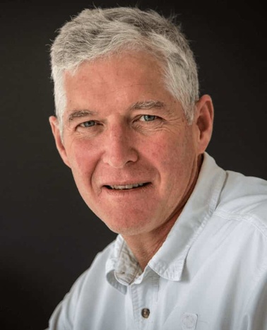 A portrait of a smiling elderly man with gray hair, wearing a white shirt against a dark background.