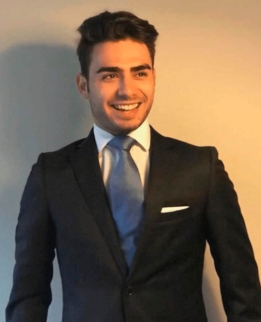 A young man smiling, wearing a dark suit with a tie and pocket square.